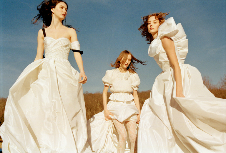Three women in flowing white dresses stand outdoors under a blue sky, looking in different directions.