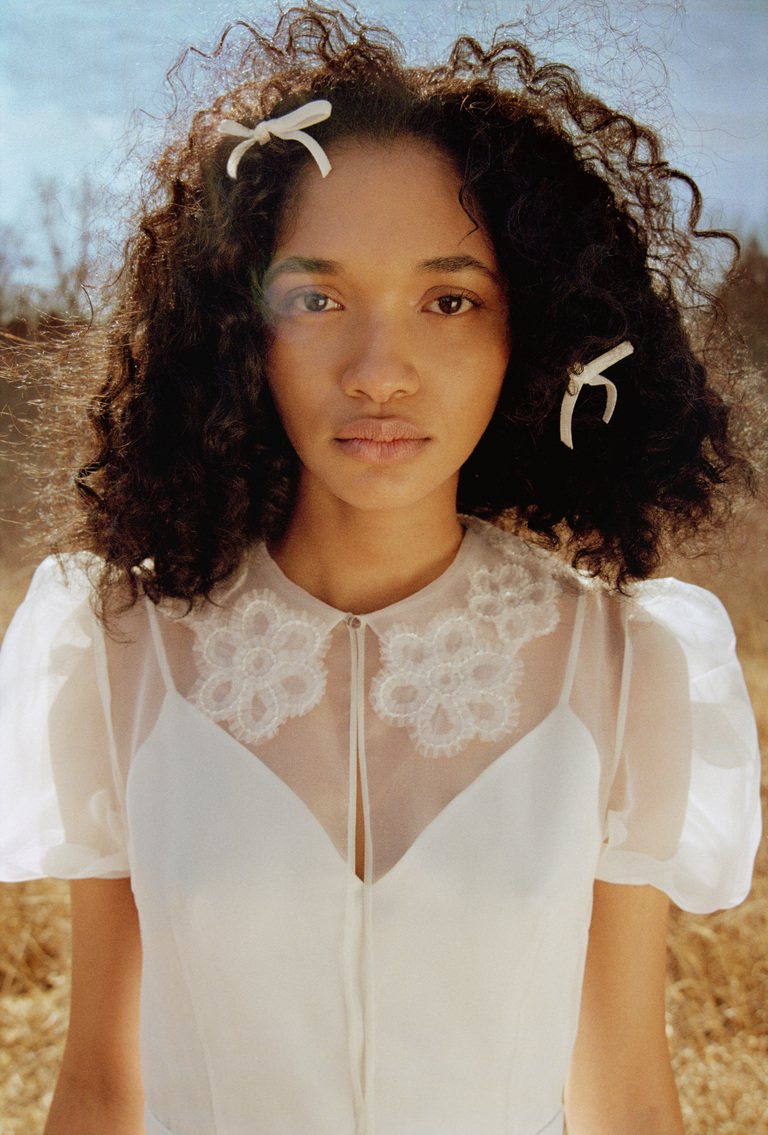 Woman in a white dress with bows in curly hair stands outdoors, looking directly at the camera.