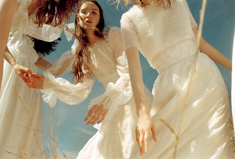 Three women in flowing white dresses stand close together outdoors under a blue sky, seen from a low angle.