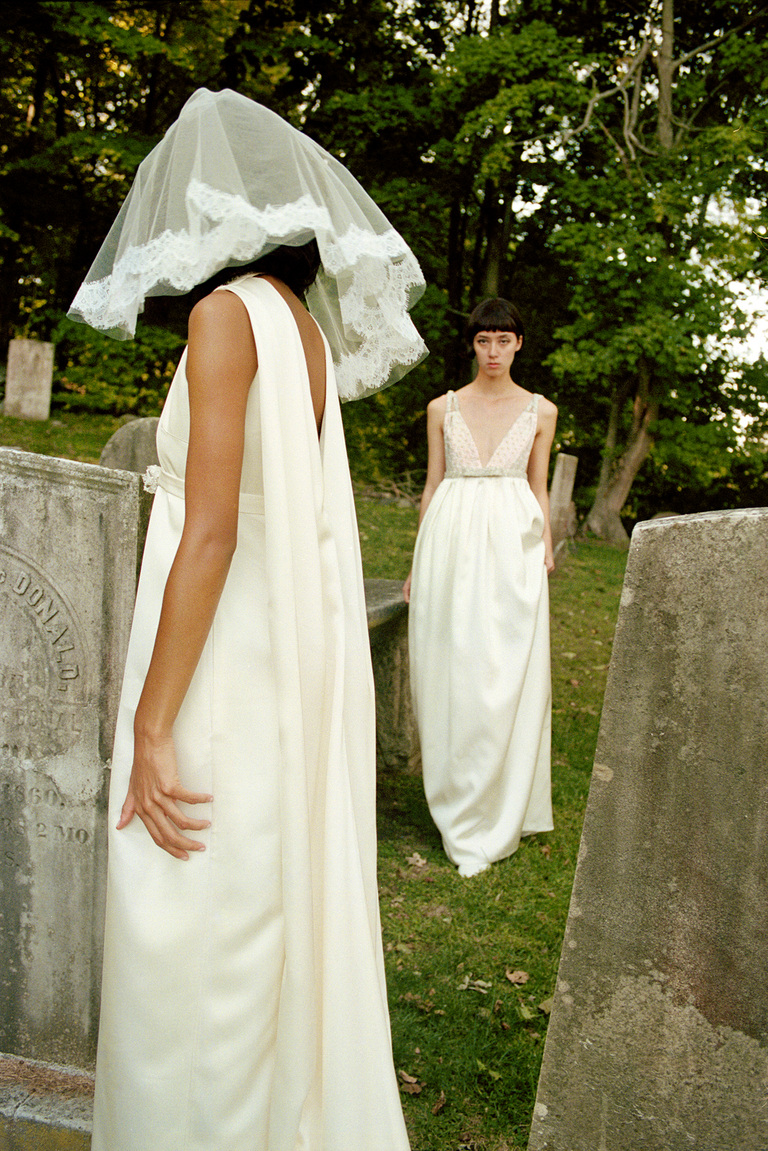 Two women in white dresses stand among tombstones; one wears a veil and faces away from the camera.