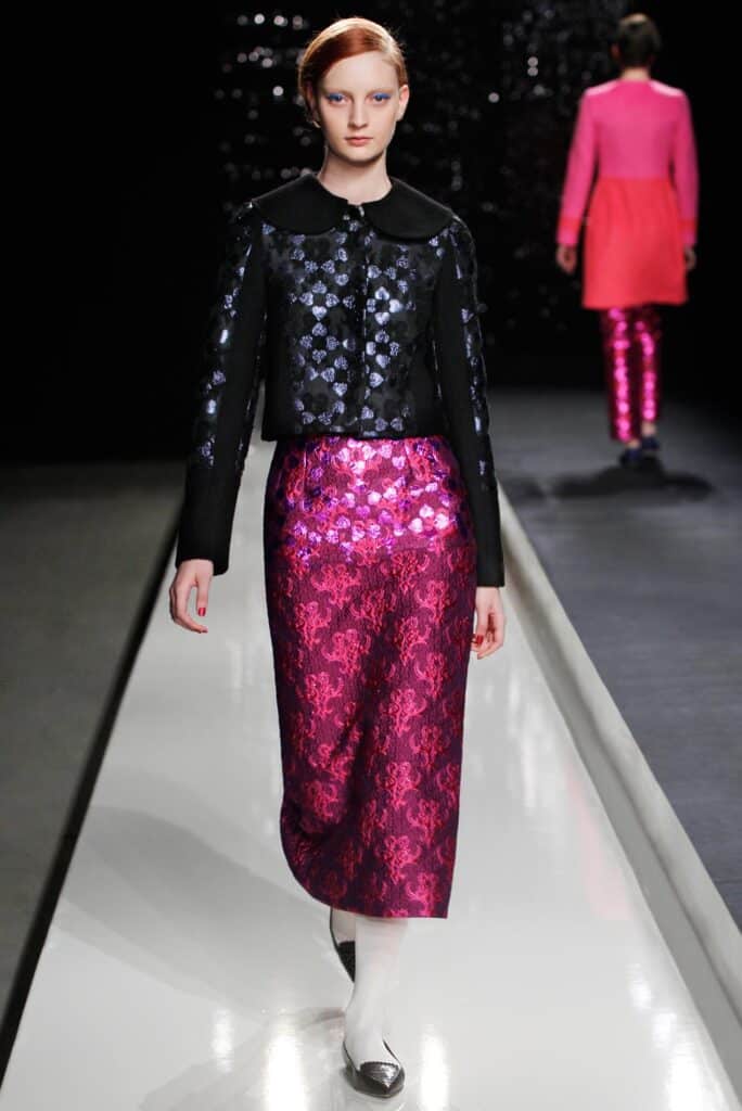 Model in a black jacket with shiny blue spots and a shiny pink skirt walks the runway.