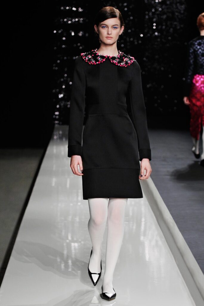 Model in a black dress with floral collar, white tights, and black heels walking on a runway.