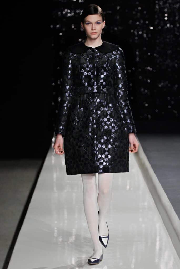A woman wearing a shiny black patterned coat, white tights, and black flats walks on a runway.