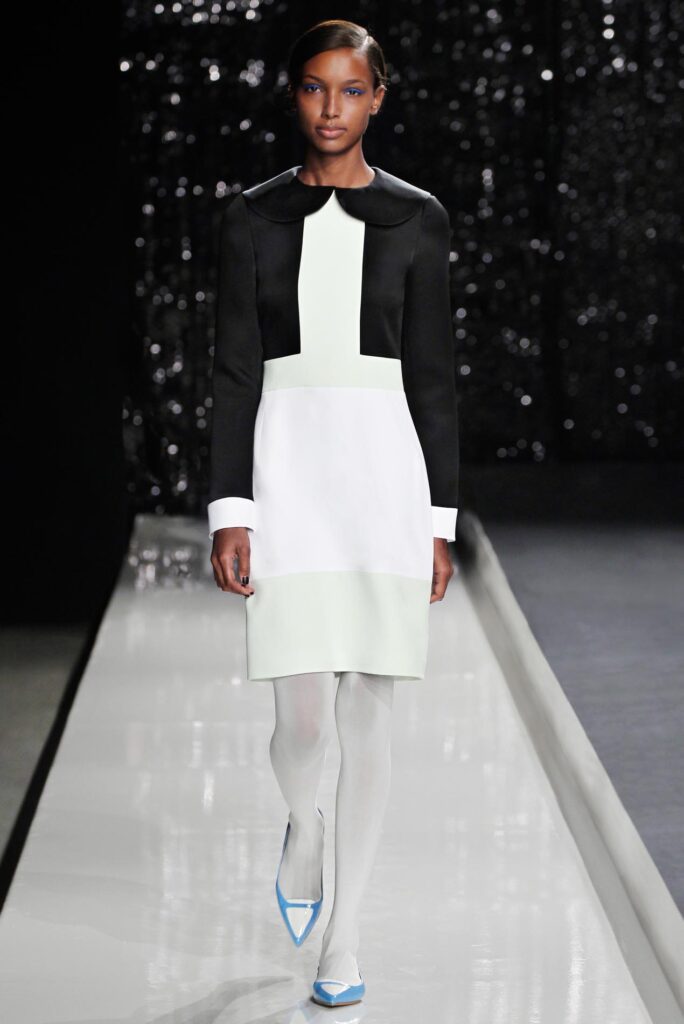Model in a black and white color-block dress with white tights and blue heels on a runway.