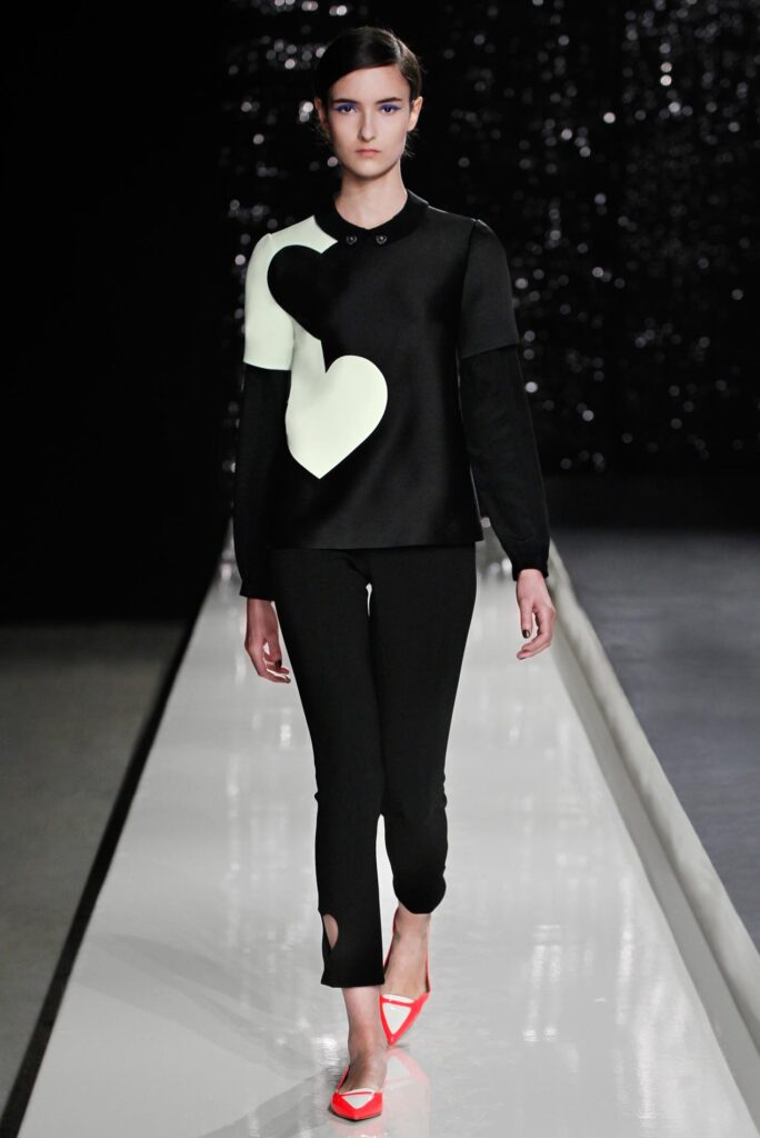 Model walks runway in black outfit with white heart shapes, black pants, and bright red pointed shoes.