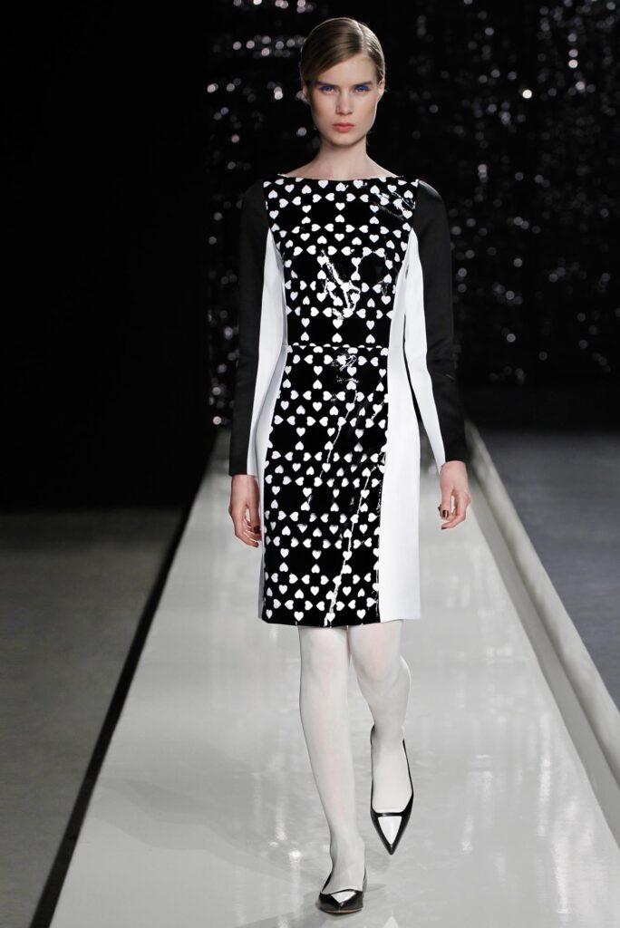 Model wears a black and white geometric dress, white tights, and black shoes on a runway with a dark backdrop.