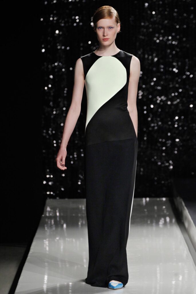 Model wearing a sleeveless black and white color-block gown walks on a runway with a glittery background.
