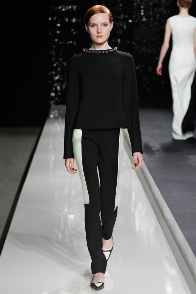 Model walks runway in black top with gem collar and black pants with white side panels, wearing pointed shoes.