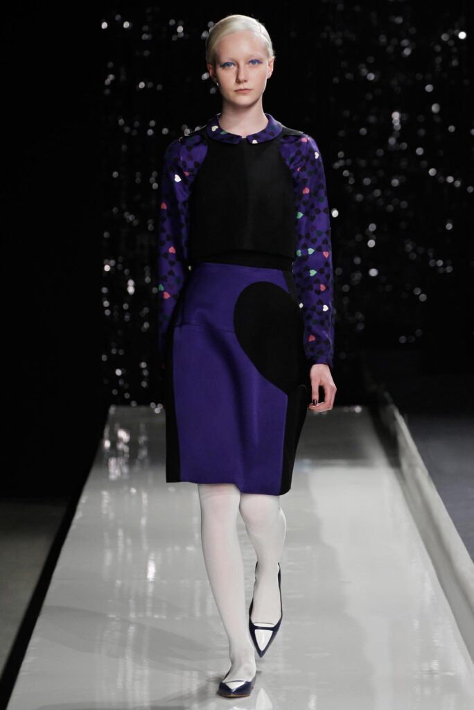 Model in a purple and black outfit with white tights walks a glossy runway, dark sparkly background behind.
