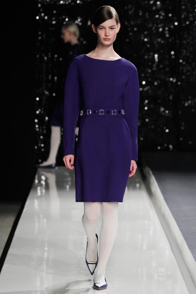 Model in a long-sleeve purple dress with a belted waist, white tights, and dark heels walks a runway.