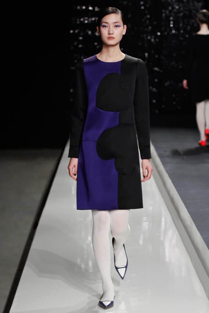 Model walks runway in a black and purple dress with heart shapes, white tights, and pointed shoes.