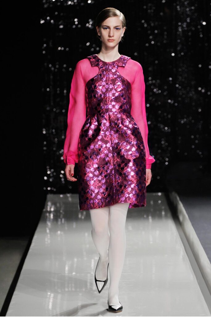 Model walks runway in shiny pink patterned dress with sheer sleeves, white tights, and black pointed heels.