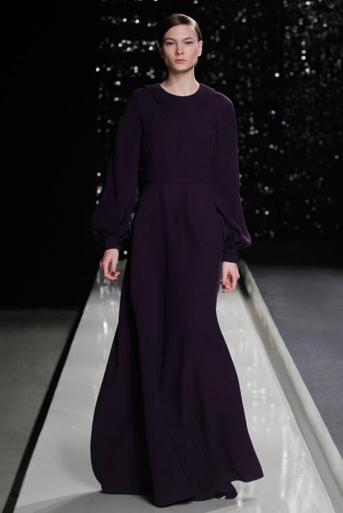 Model in a long-sleeved, dark purple gown walking down a runway with a black sequin background.