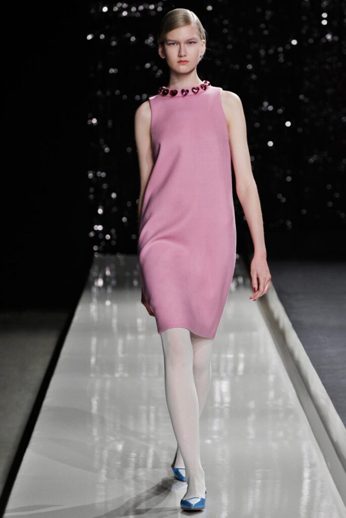 Model in a pink sleeveless dress with heart necklace, white tights, and blue shoes walks on a runway.