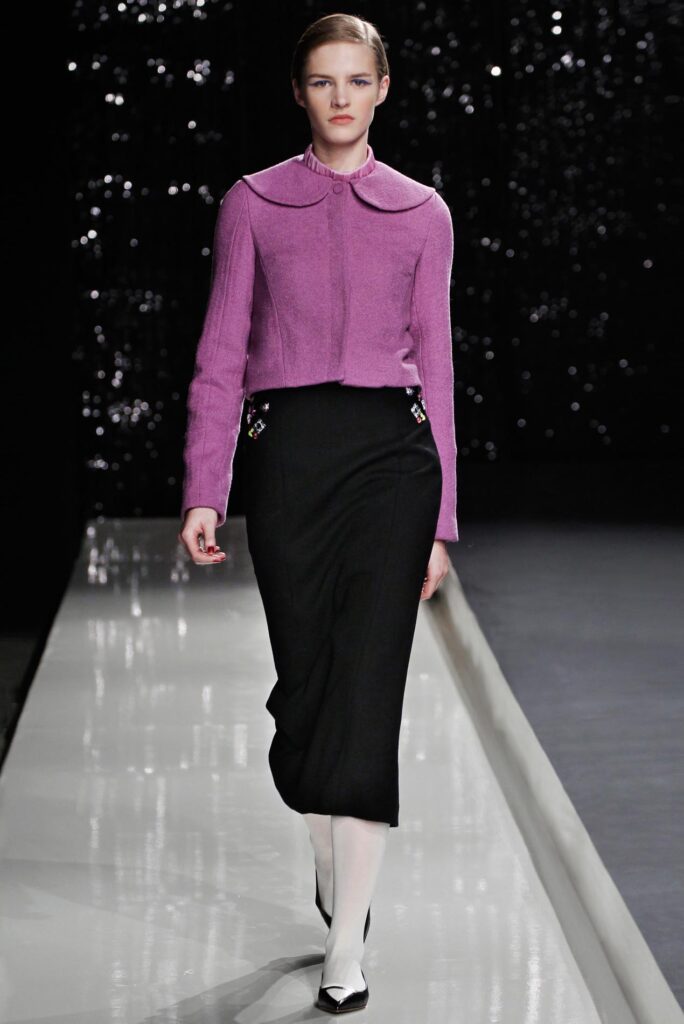 Model walks runway in a pink jacket, black skirt, white tights, and heels against a dark, sparkly background.