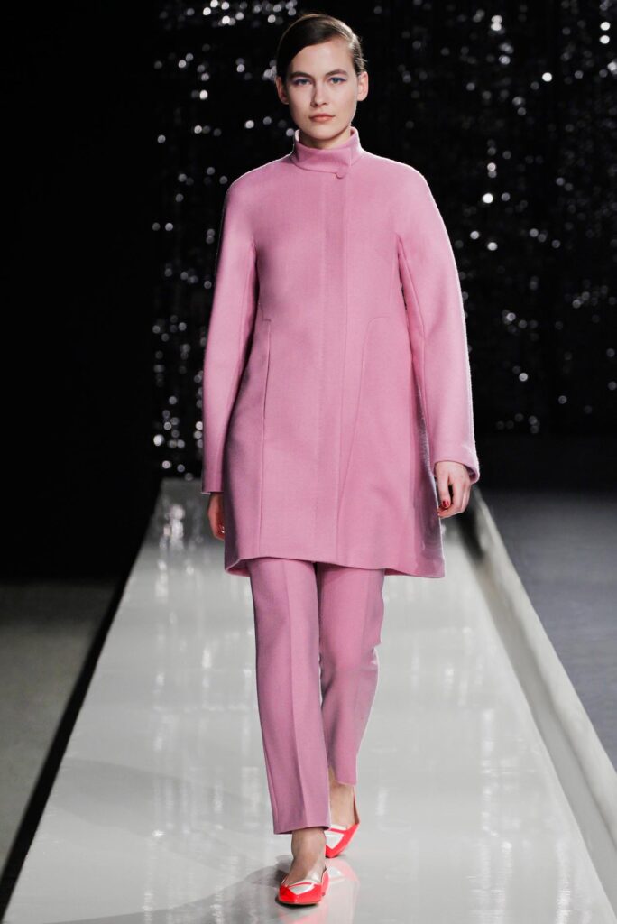 Model in a pink coat and pants walks the runway; background is dark with sparkling lights.