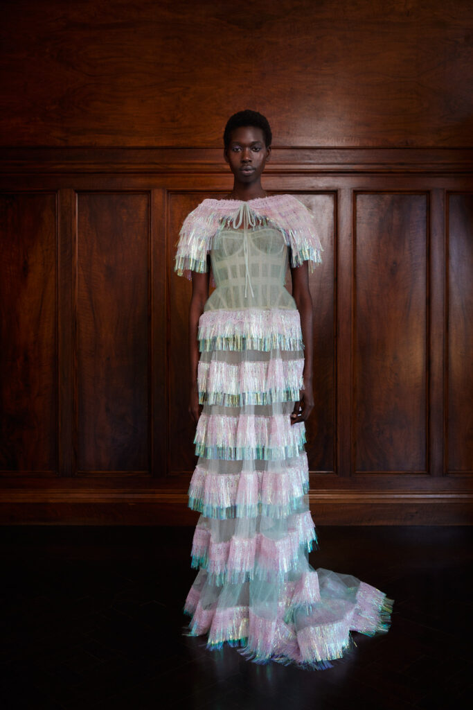 A woman in a pastel, tiered, and fringed gown stands in front of paneled wooden walls.