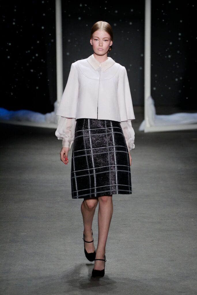 Model wearing a white blouse and black plaid skirt walks on a runway with a starry background.