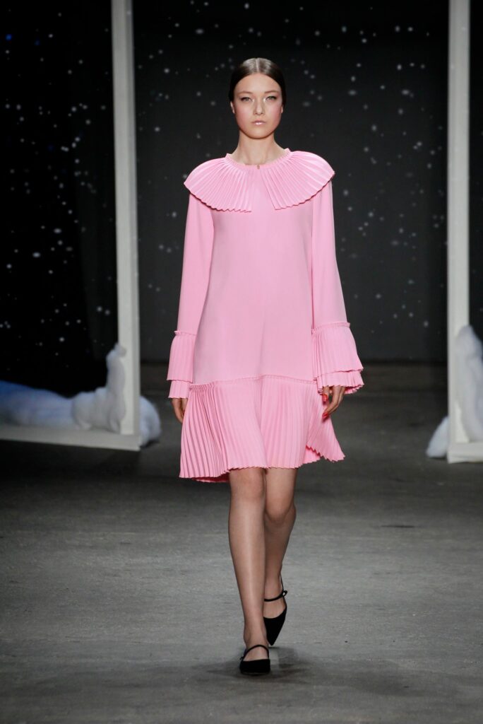 Model walks runway in a pink pleated dress with ruffled collar, black shoes, and hair slicked back.