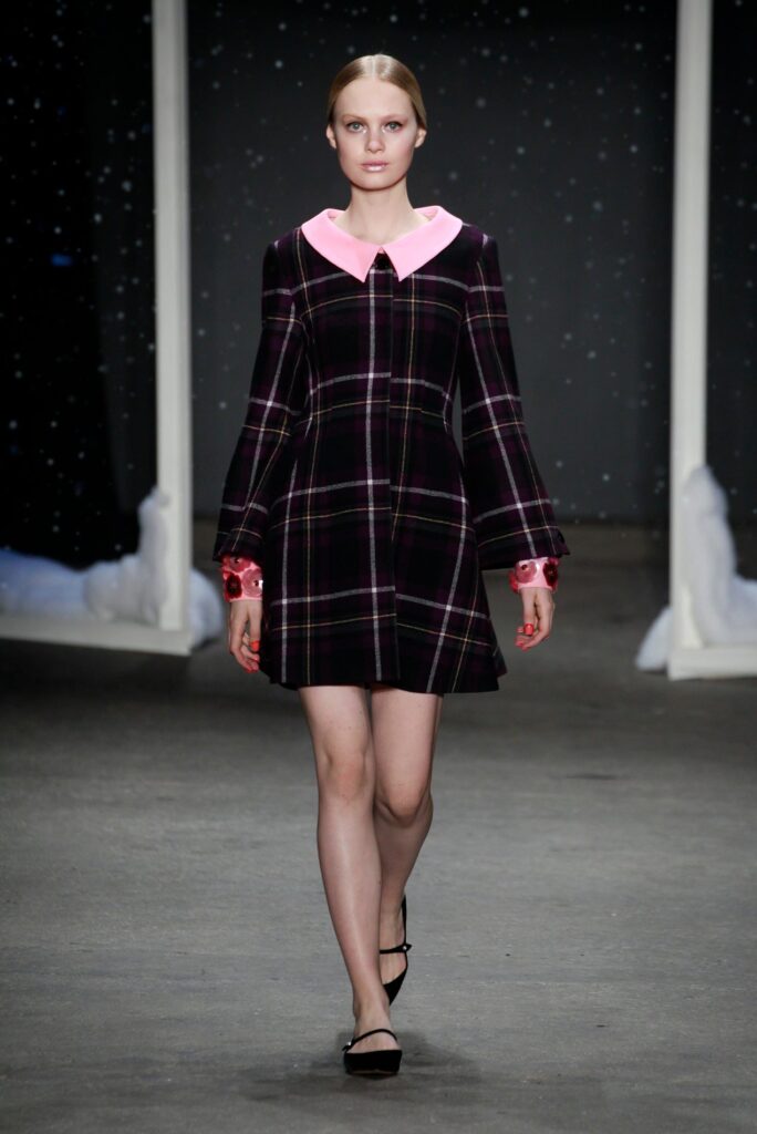 Model walks runway in a plaid dress with a pink collar and red cuff details, black flats, starry backdrop.