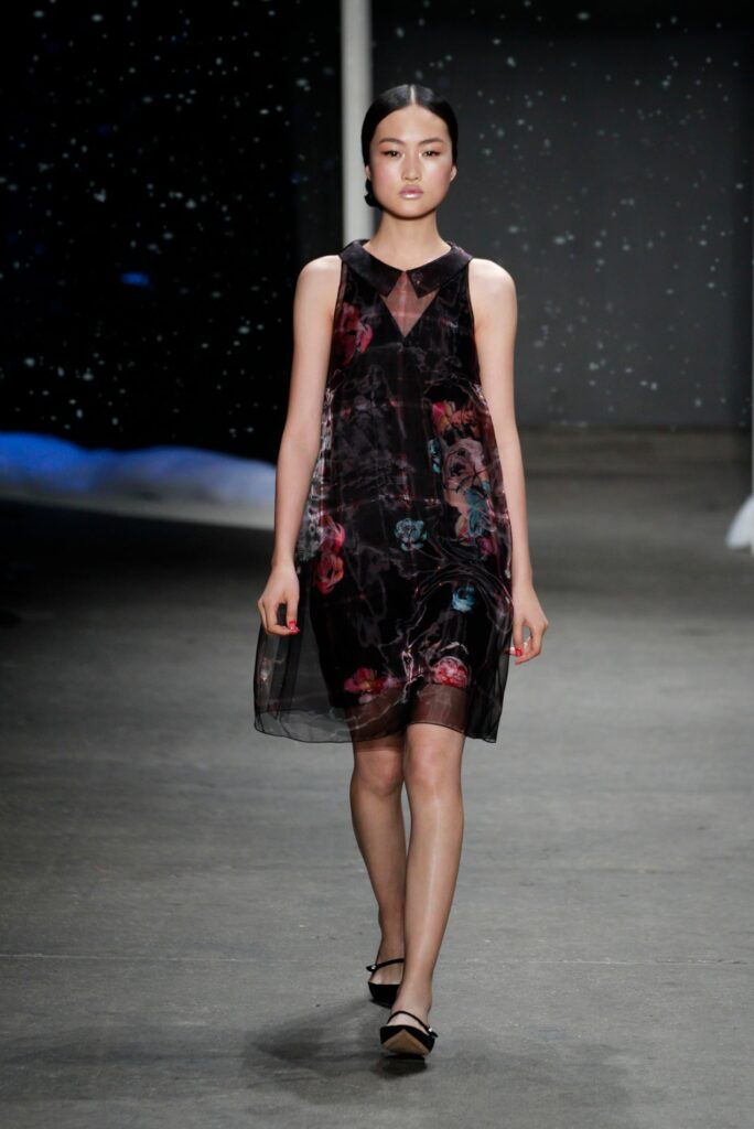 Model walks on runway in a sleeveless black dress with floral patterns and black flats. Dark, starry backdrop.
