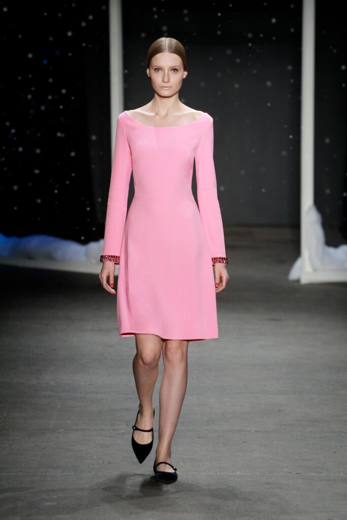 Model in a long-sleeve pink dress and black shoes walks on a runway with a dark, starry background.
