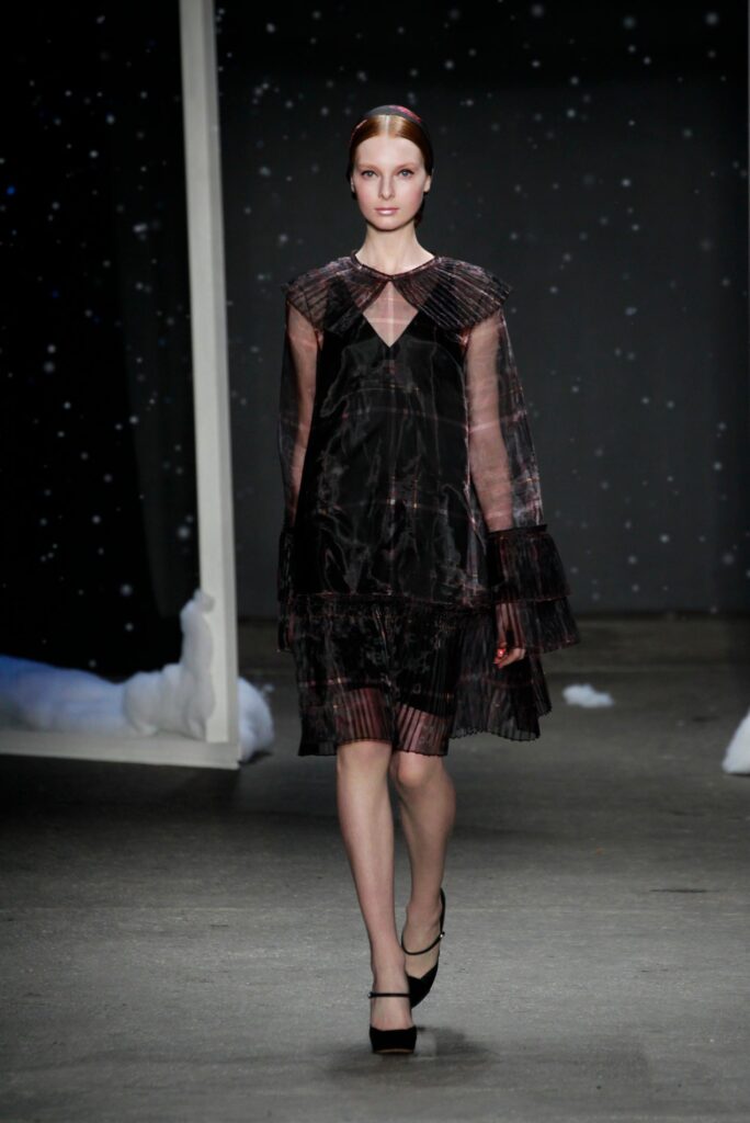 Model in a sheer black dress walks a runway with a snowy night backdrop and black heels.