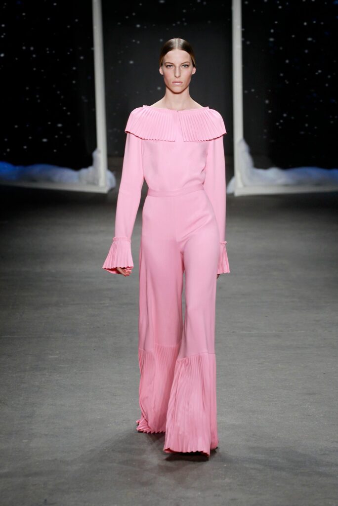 Model walks runway in a pink jumpsuit with pleated collar, cuffs, and pant legs, set against dark background.
