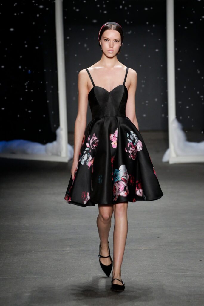 Model in a black floral dress with thin straps walks on a runway, background features a night sky theme.