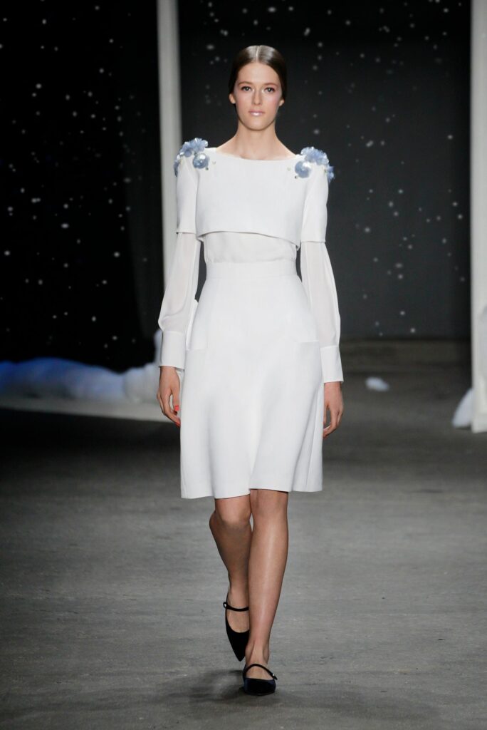 Model in a white dress with sheer sleeves and floral shoulder details walks on a runway with a dark backdrop.