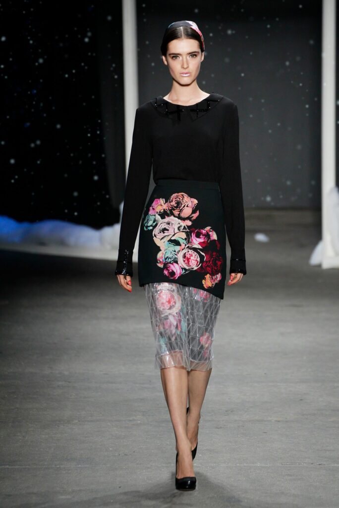 Model walks runway in black top, floral skirt, sheer overlay, and black heels; starry backdrop behind.