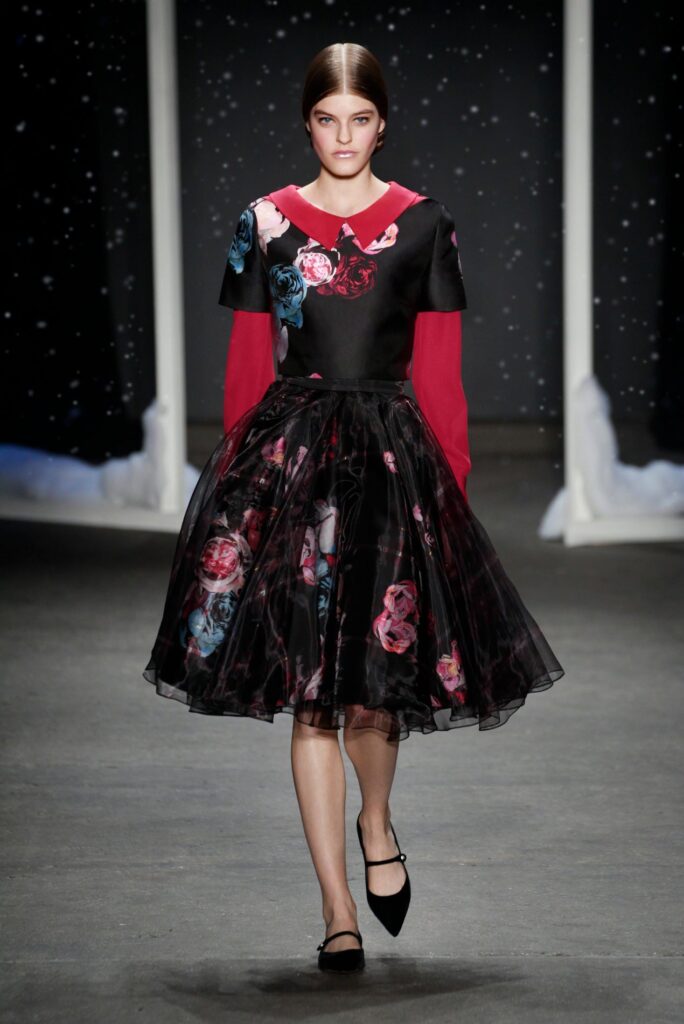 Model in a black floral dress with red sleeves and collar walking on a runway with a dark, starry backdrop.