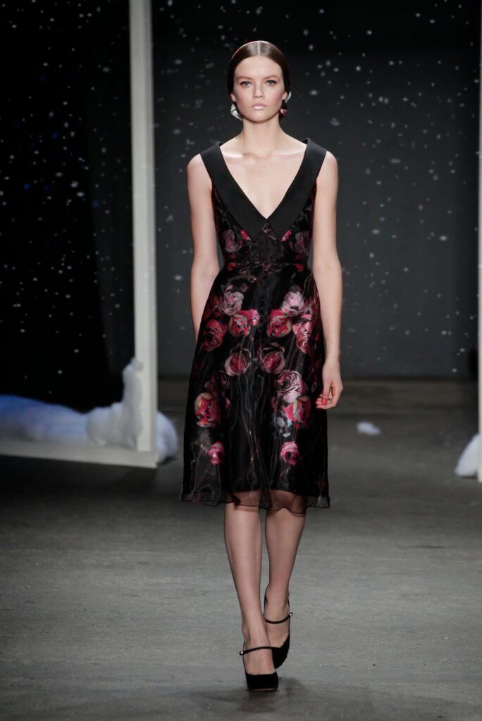 Model walks runway in black dress with red floral print, background features dark sky with white specks.