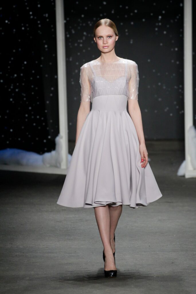 Model in a light grey, knee-length dress with sheer sleeves walks on a dark runway with star-like lights.