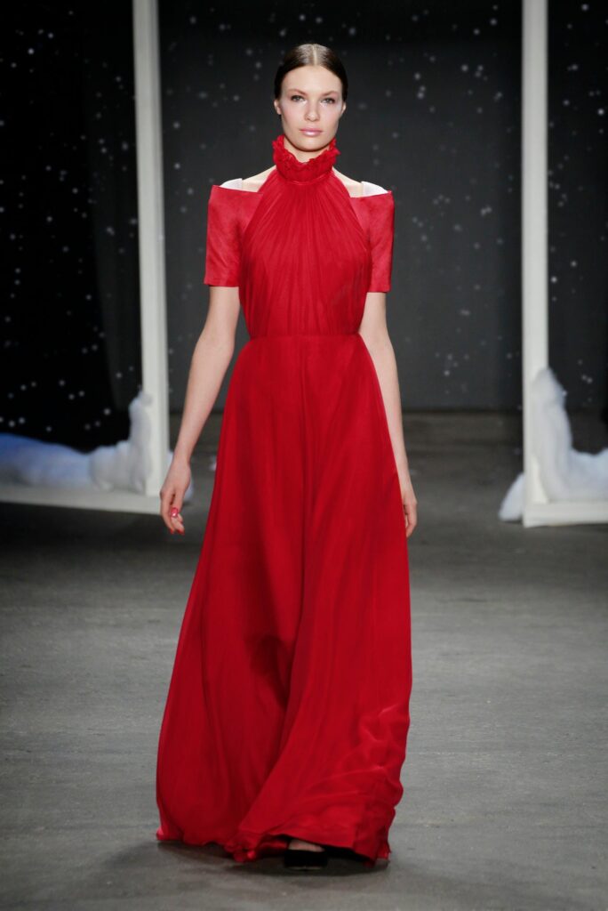 Model in a flowing red gown with cut-out shoulders walking on a runway with a dark, starry background.
