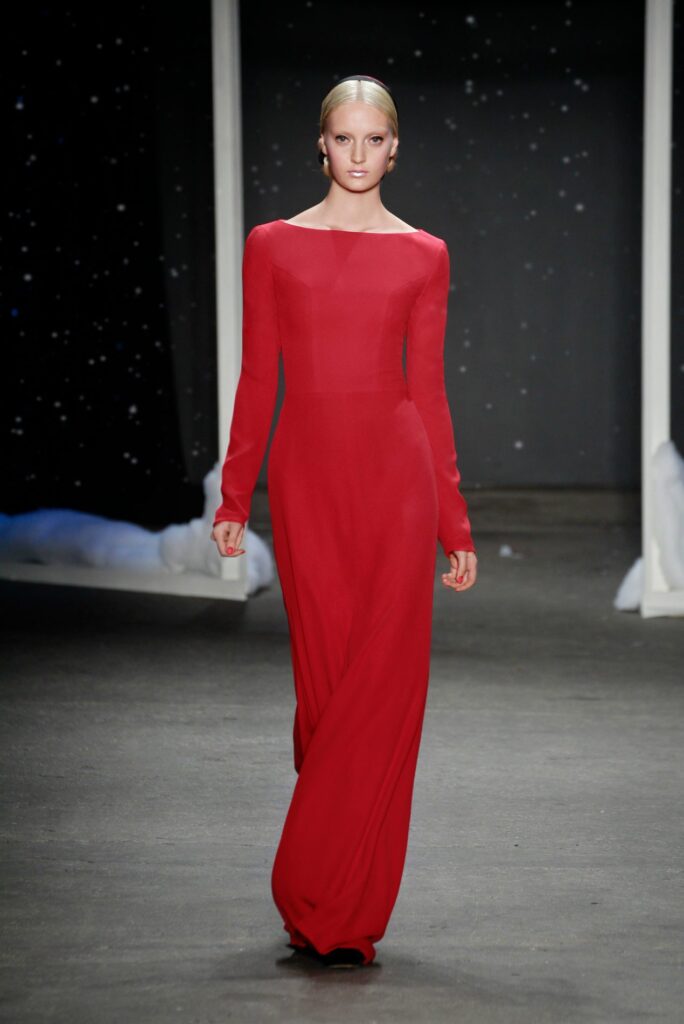 Model in a long-sleeved red gown walks down a runway with a dark, starry background.