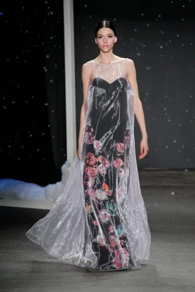 Model in a black floral gown with a sheer overlay walks on a runway, starry backdrop behind her.