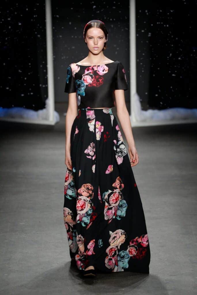 Model walks runway in a black floral gown with short sleeves and a full skirt against a starry backdrop.
