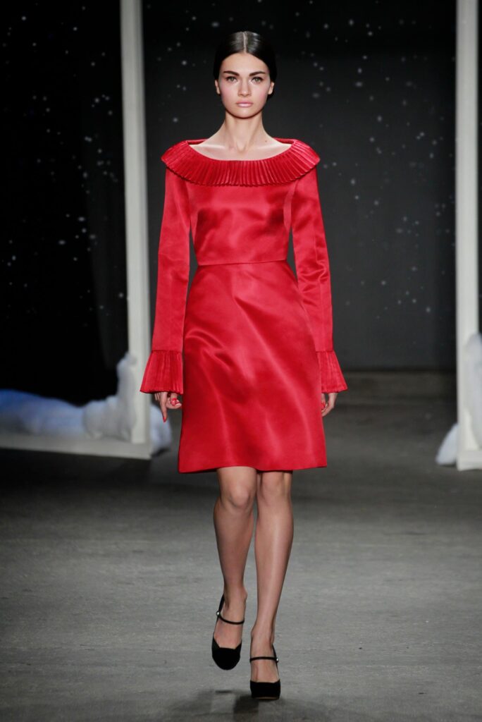 Model in a red long-sleeve dress with flared cuffs walking on a runway, black heels, dark background.