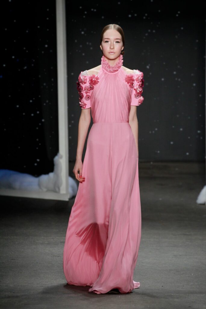 Model in a flowing pink gown with floral appliqués on the shoulders, walking on a runway.