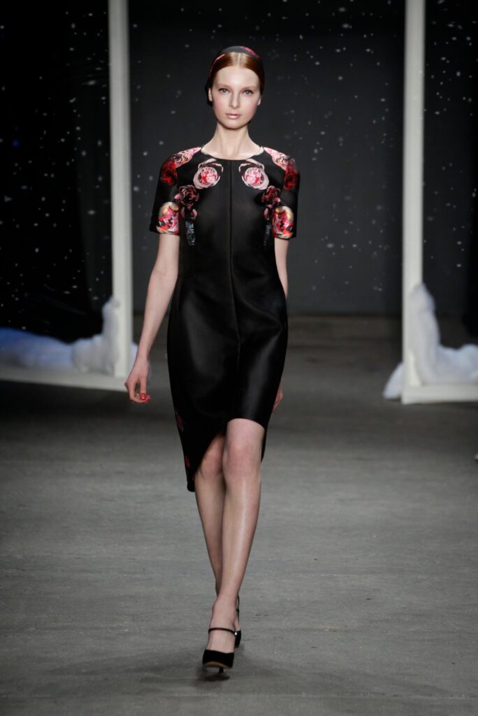 Model walks runway in black dress with red floral details, black heels, dark backdrop with white specks.