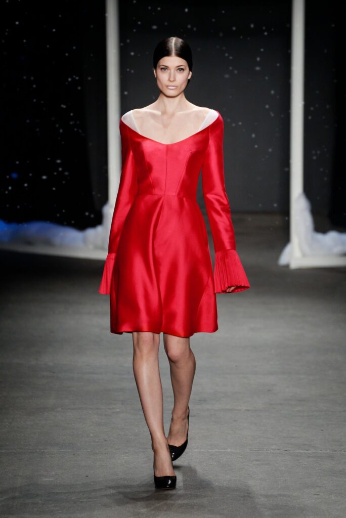 Model in a red, long-sleeved dress with pleated cuffs walks on a runway with a starry background.