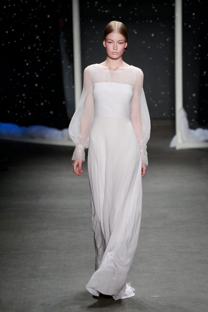 Model in a long white dress with sheer sleeves walks down a runway with a dark starry background.