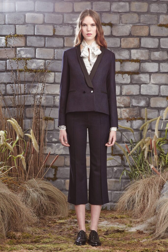 Woman in dark suit with ruffled white shirt stands in front of stone wall, surrounded by tall grass.