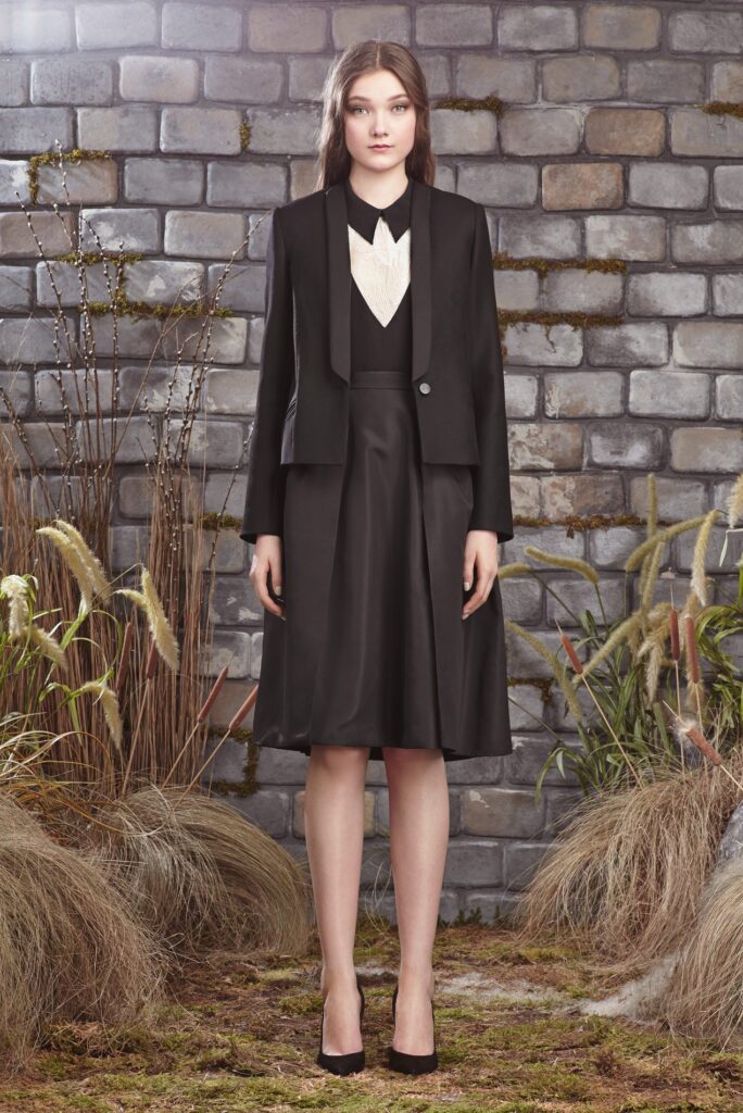 Woman in a black skirt suit with a white blouse stands in front of a brick wall and plants.