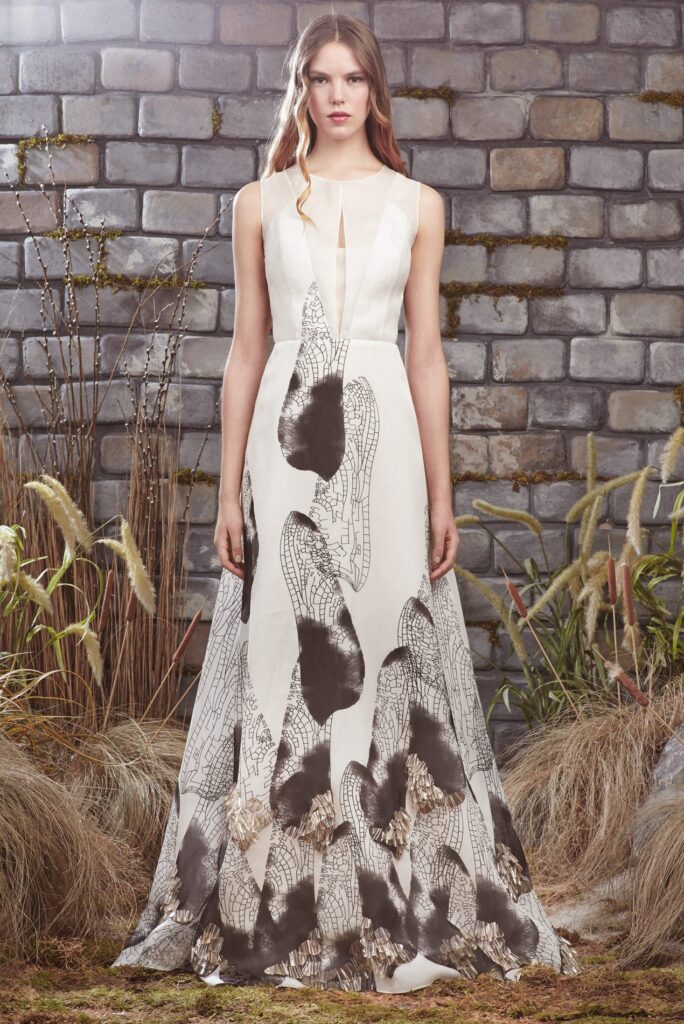 Woman in a sleeveless white dress with black abstract patterns stands outdoors by a stone wall and tall grasses.