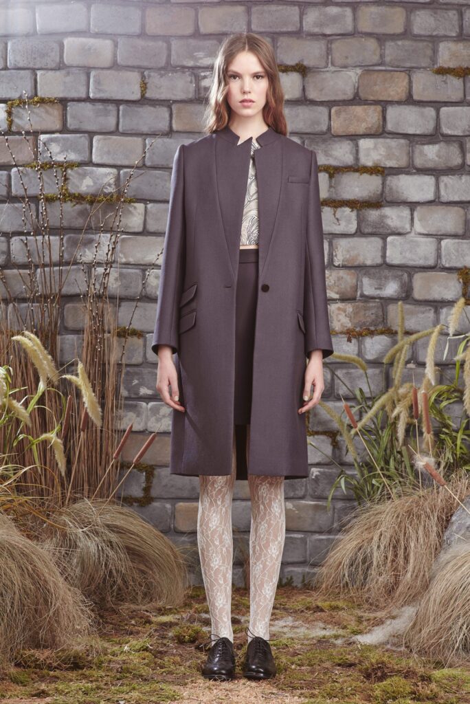 Woman in a gray coat, lace tights, and black shoes stands in front of a brick wall and tall grasses.