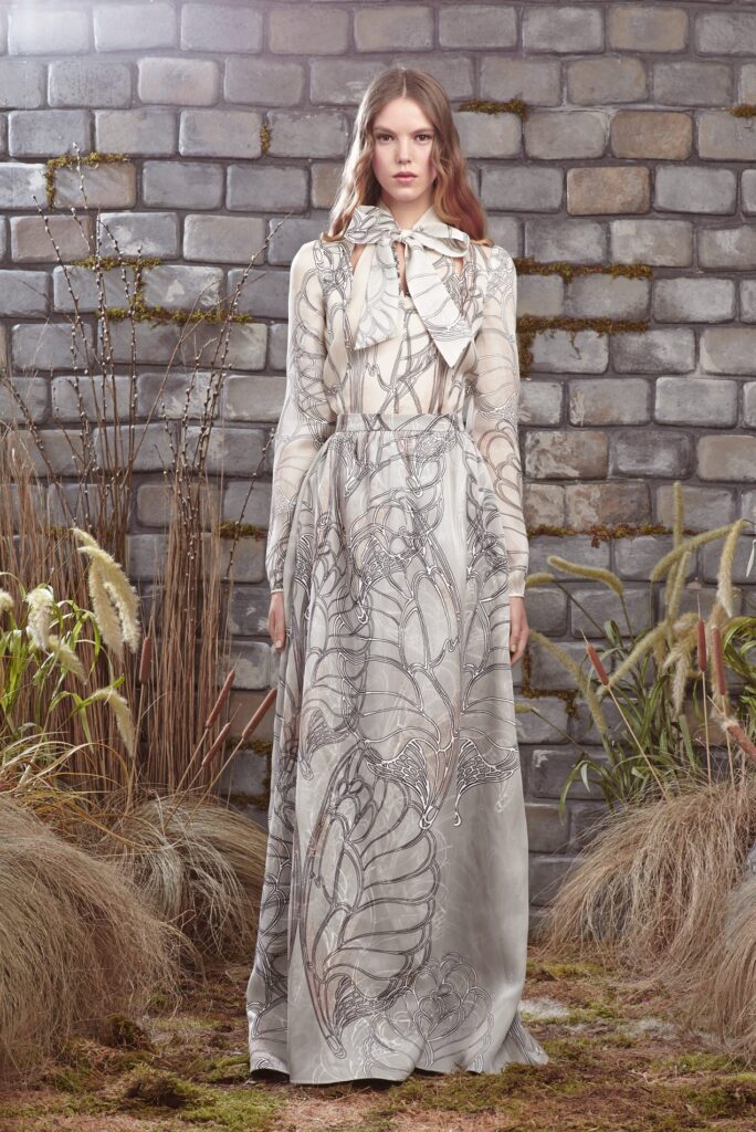 Woman in a long, patterned gown stands against a stone wall with dry grass and plants around her.