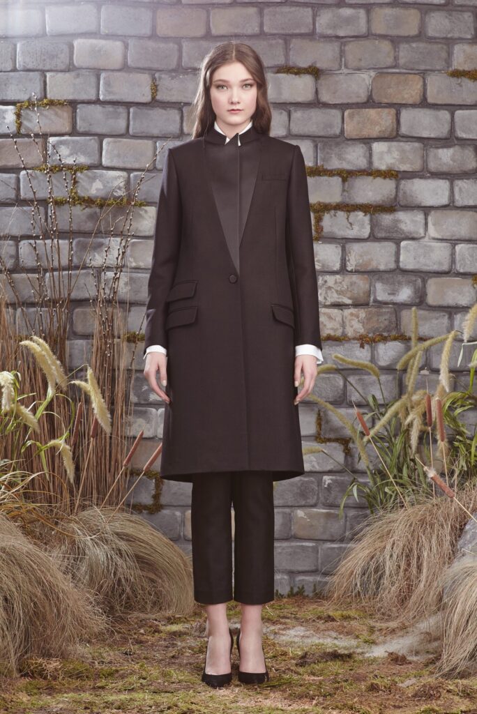 Woman in a black suit and coat stands on moss, in front of a brick wall with plants around her.
