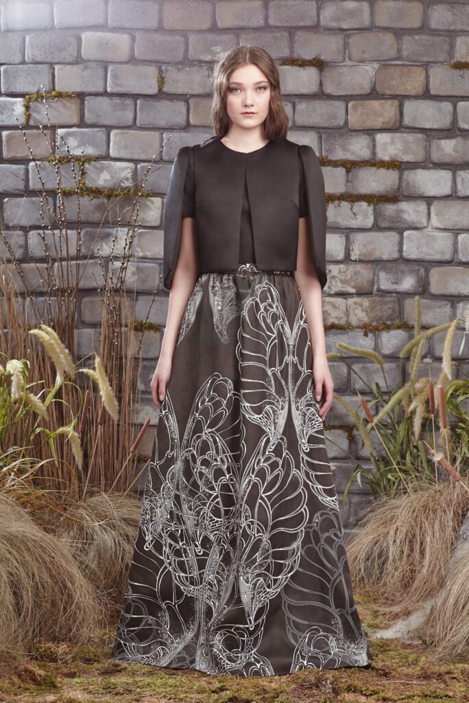 Woman in black top and long skirt with white abstract patterns, standing by a stone wall with tall grass.